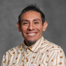 Man with short, dark hair and chin patch wearing tan shirt with brown and gold print